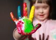A young girl - not in focus but smiling - stretches out her hand to camera: her fingers and palm are daubed with patches of different coloured paint