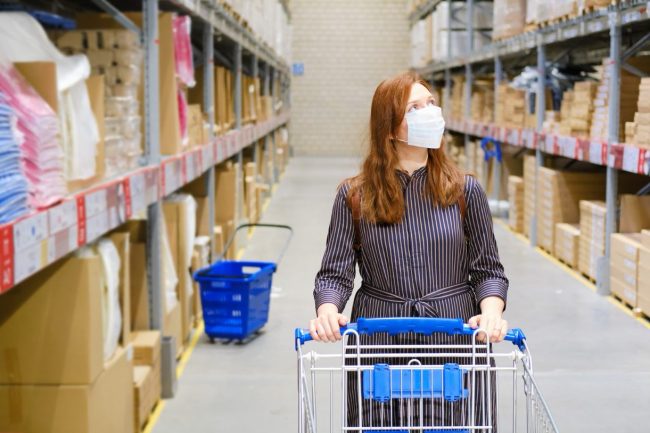 A woman wearing a medical mask and pushing a shopping trolley in warehouse style shop with big cardboard boxes of goods stacked on tall shelves