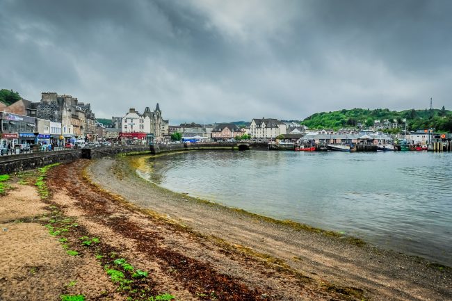 View from a beach looking towards the buildings of the Scottish town of Oban including shops and cafes, cars in the streets and boats in the harbour. There is a hill beyond the town with a phone mast.